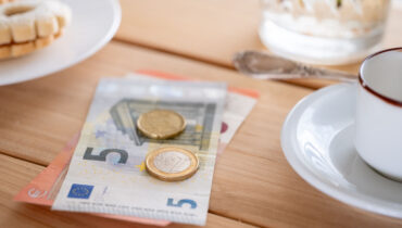Euro banknotes and coins on wooden table with cookie and coffee cup nearby casual dining setting, Euro currency placed beside snack and coffee suggesting small purchase or tip, hospitality